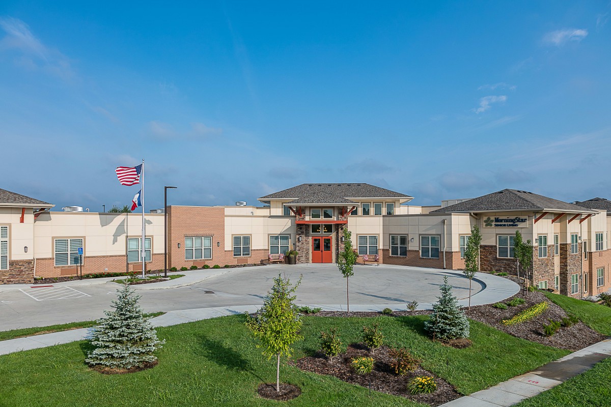 A modern school building with a circular driveway, landscaped grounds, and a flagpole against a clear blue sky.