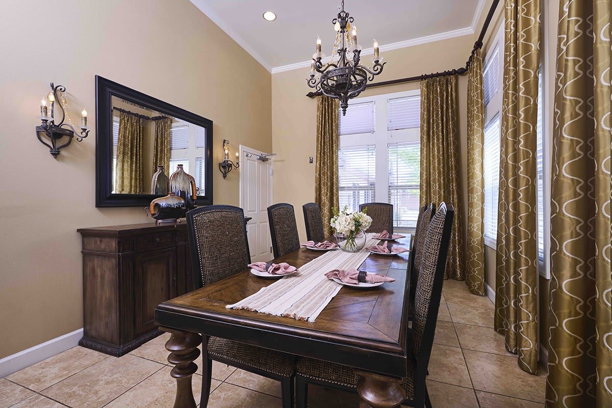 A classic dining room featuring a wooden dining table with six chairs, an elegant chandelier, and large windows with curtains.