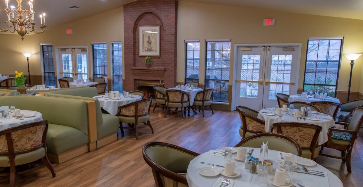 An elegant dining area with tables and chairs set for a meal, featuring large windows and warm lighting.