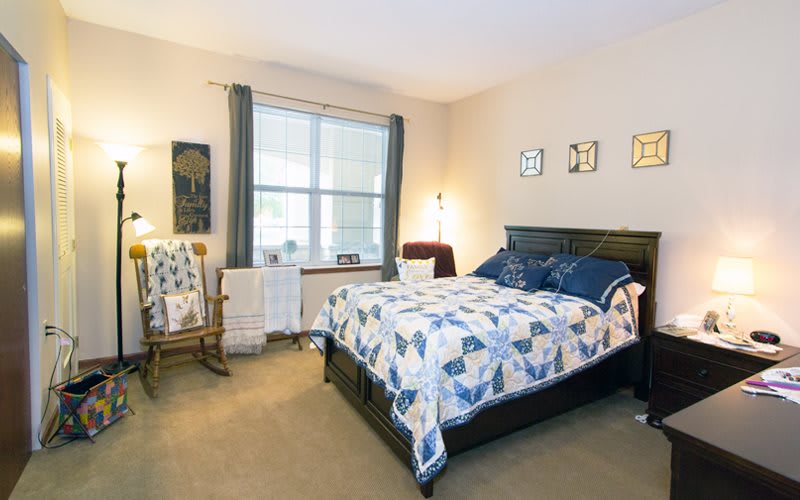 A cozy bedroom with a neatly made bed featuring a patterned quilt, a large window with curtains, and a rocking chair in the corner.