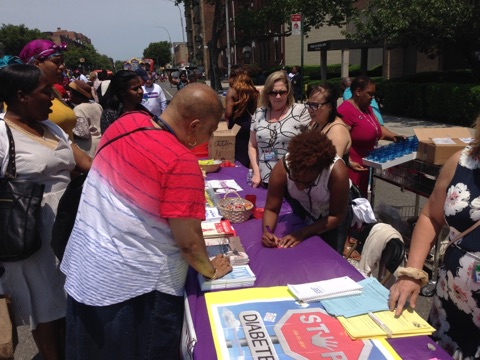 A lively community event with people engaging at an information booth under the summer sun.