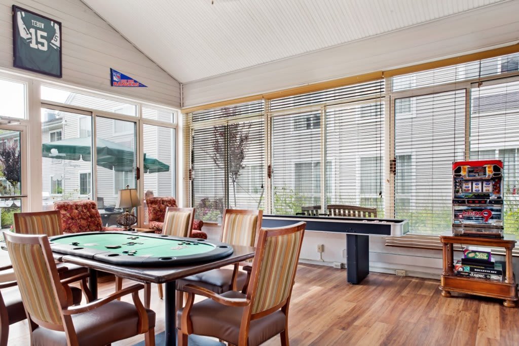 A bright indoor game room featuring a poker table, chairs, and a foosball table with large windows bringing in natural light.