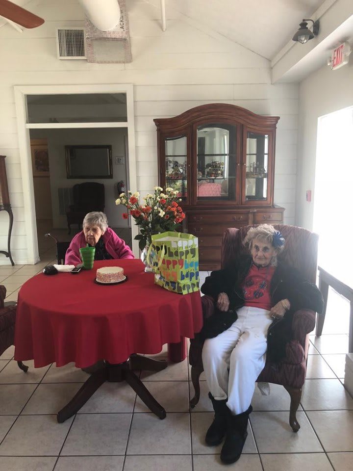 Two elderly individuals celebrating a birthday with a cake, flowers, and gift inside a home.