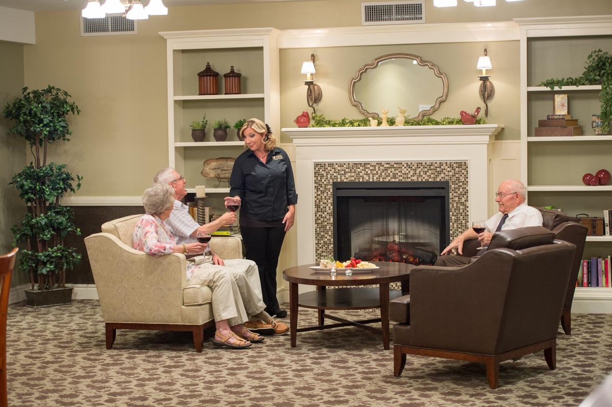 A group of elderly individuals sitting in a cozy living room, engaged in conversation near a fireplace.