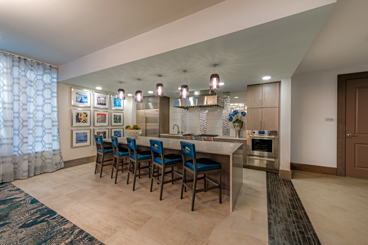 A modern kitchen with a large island, blue bar stools, and pendant lighting, featuring artwork on the walls.