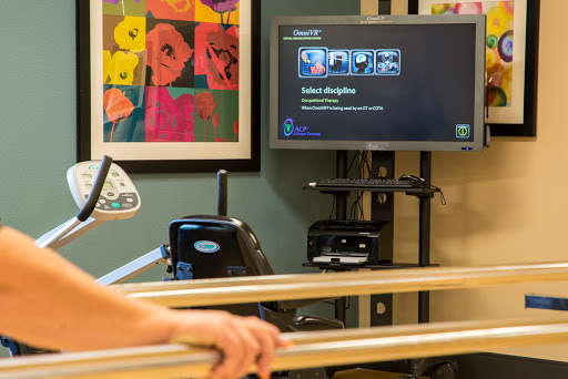 A therapy room featuring exercise equipment and a monitor displaying information, complemented by colorful artwork on the wall.