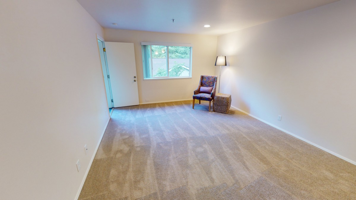 A bright, empty living room with carpet flooring, featuring a window, a chair, and a floor lamp.