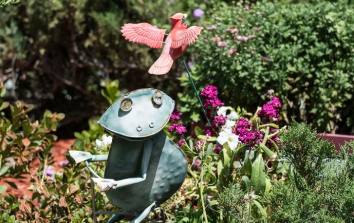 A whimsical garden scene featuring a metal frog sculpture among vibrant flowers, with a red metal bird flying overhead.