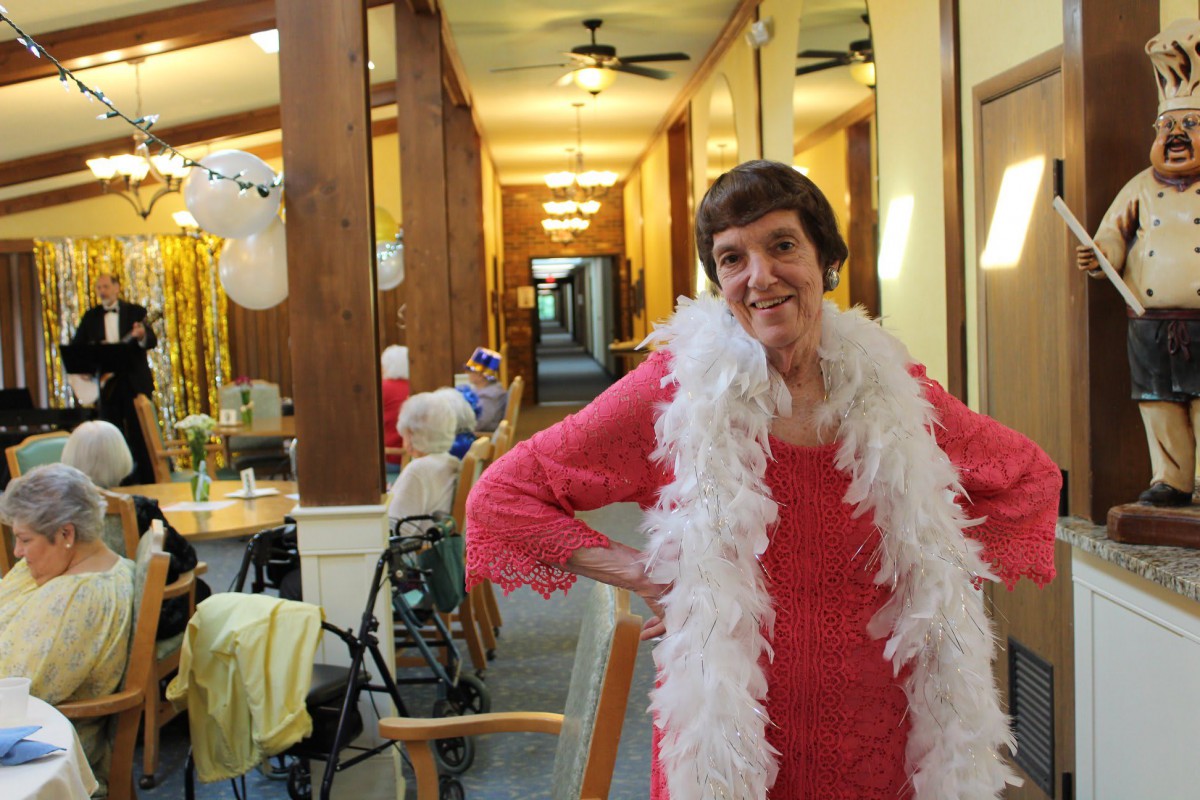 An elderly woman dressed in a red outfit and white boa poses happily in a nursing home hallway.