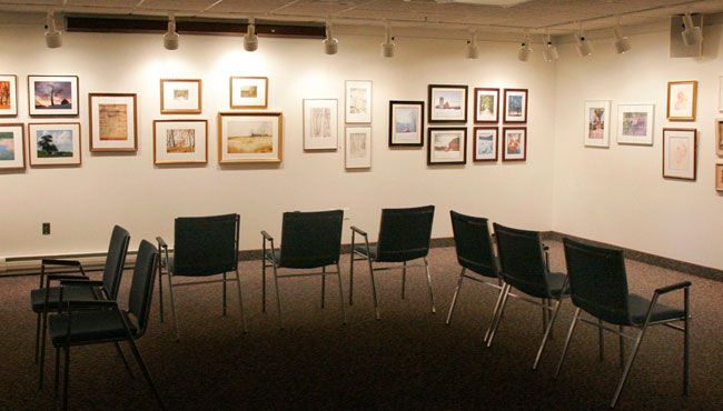 A quiet art gallery room with paintings on the wall and chairs arranged for visitors.