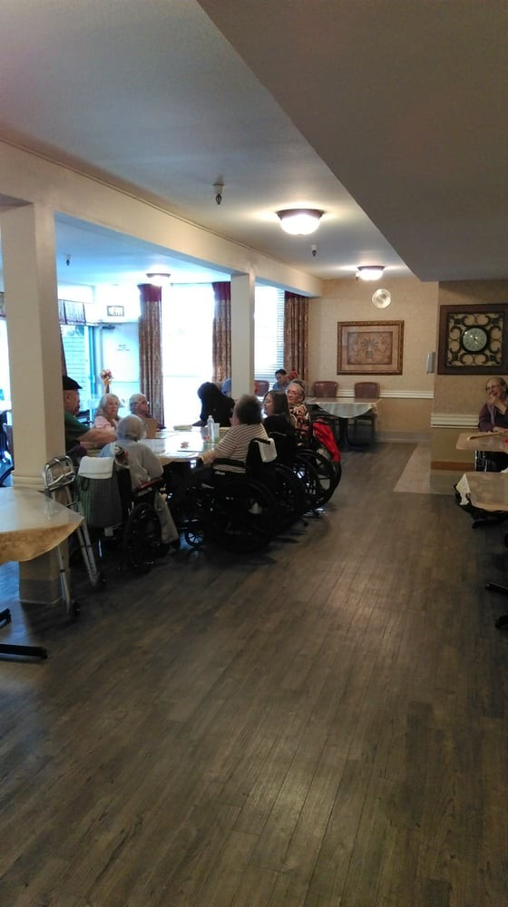 A group of elderly people gather in a community room, some using wheelchairs, during the day.
