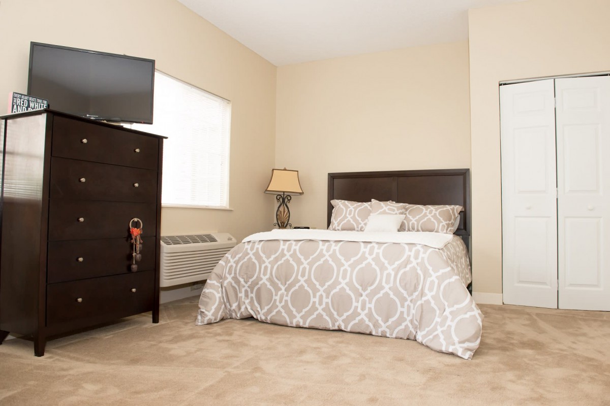 A cozy bedroom with neutral tones, featuring a bed, dresser, lamp, and mounted TV.