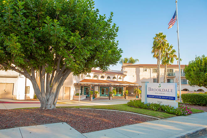 A building with a prominent tree in front and a sign at the entrance, surrounded by palm trees on a clear day.