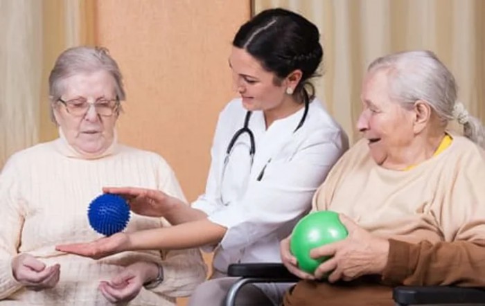 A healthcare professional assists elderly individuals with an exercise activity using colored balls in a nursing home setting.