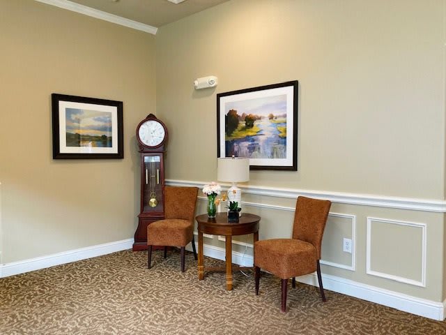 A cozy corner of a living room featuring two chairs, a small table with flowers and a lamp, a grandfather clock, and framed artwork on the walls.