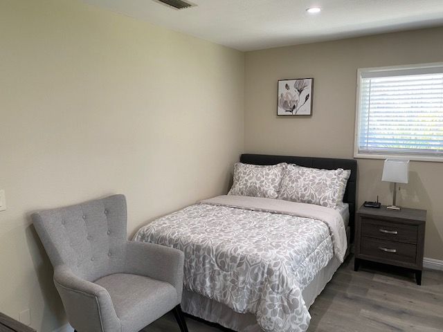 A cozy bedroom with a neatly made bed, a gray chair, and a small nightstand by the window.
