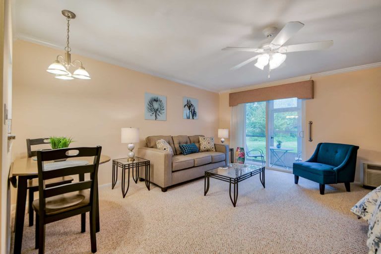 A cozy living room with a sofa, tables, and ceiling fan, featuring a dining area and large windows.