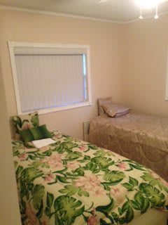 A small bedroom featuring two single beds, one with a floral bedspread, and a window with closed blinds.