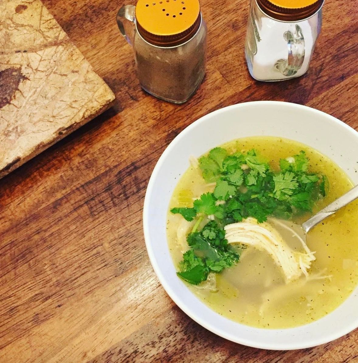 A warm bowl of chicken soup garnished with fresh cilantro, served with bread and spices on a wooden table.