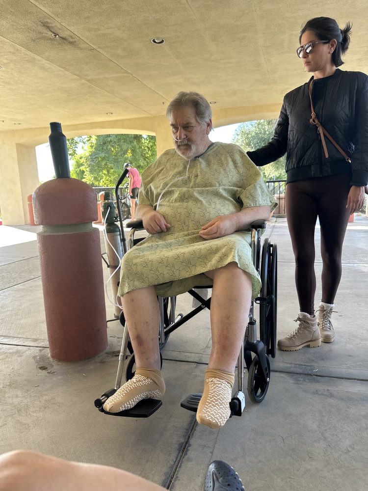 An elderly person in a wheelchair wearing a hospital gown, accompanied by a caregiver outdoors.