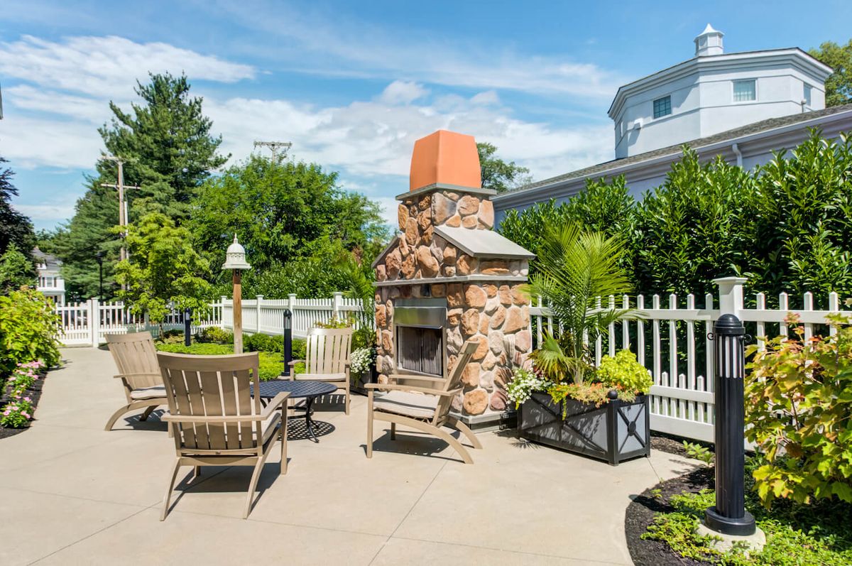 A sunny outdoor patio featuring a stone fireplace, surrounded by chairs and lush greenery.