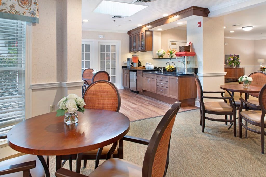 A cozy dining area with round wooden tables and chairs, featuring a kitchenette in the background.