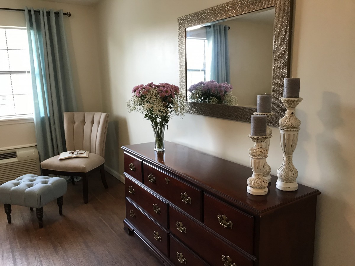 A well-decorated bedroom corner featuring a wooden dresser with a mirror, a vase of flowers, decorative candles, and an upholstered chair.