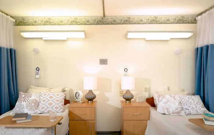 A clean and well-lit hospital room with two beds and nightstands equipped with lamps.