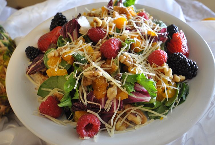 A colorful fruit and nut salad with mixed greens, topped with raspberries, blackberries, mandarin oranges, walnuts, and shredded cheese.