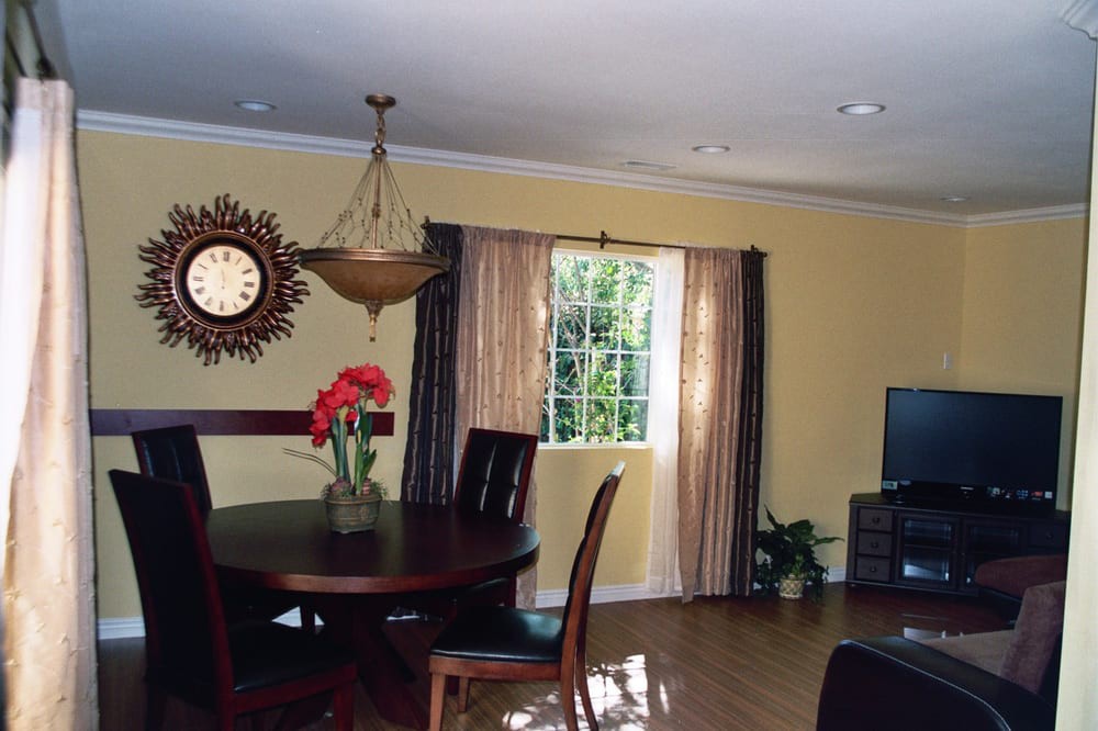 A bright living and dining area with a wooden table and chairs, a decorative clock on the wall, and sunlight streaming through the window.