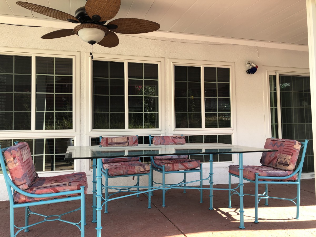 An outdoor patio area featuring a glass table with metal chairs and a ceiling fan above, situated in front of multiple large windows.