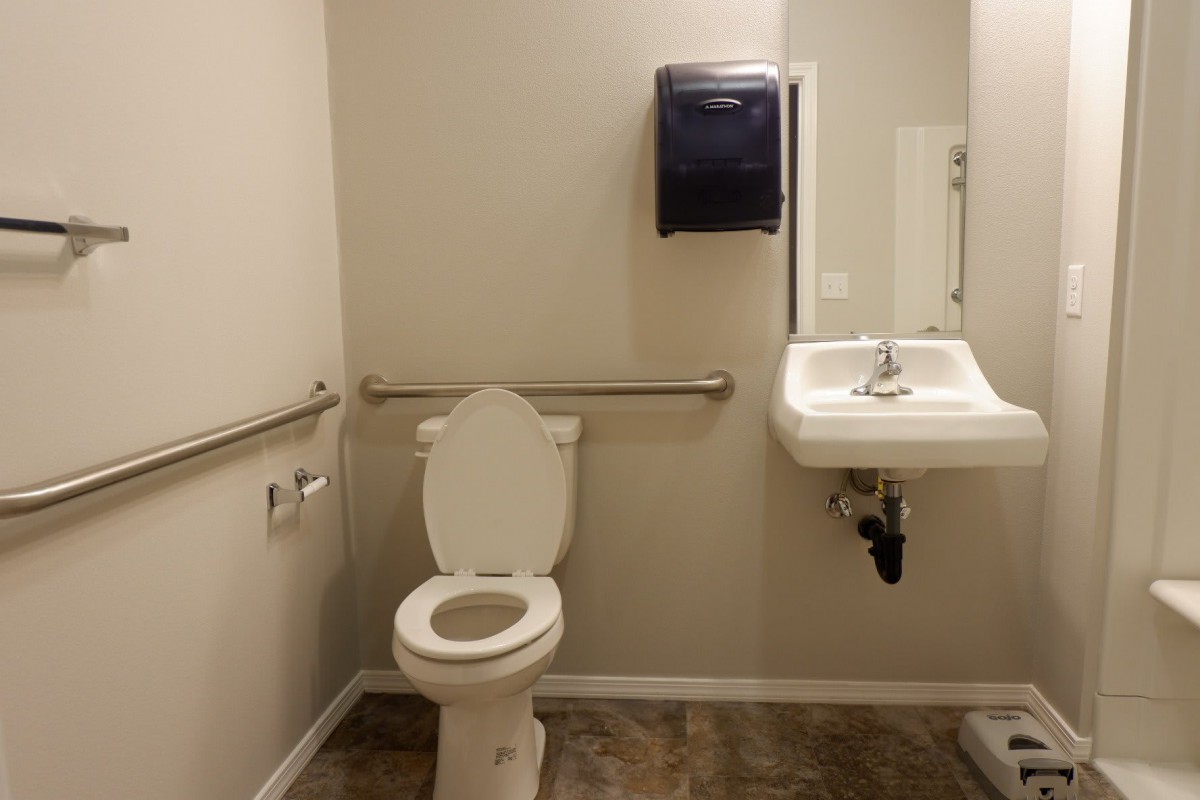 A clean and accessible public restroom featuring a toilet, sink, mirror, and handrails.