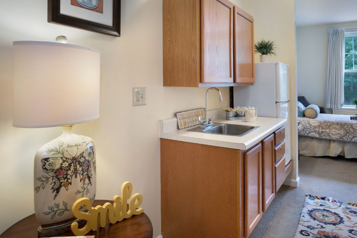 A compact kitchen area with wooden cabinets, a sink, and a refrigerator, accented by a decorative lamp and 'Smile' sign.