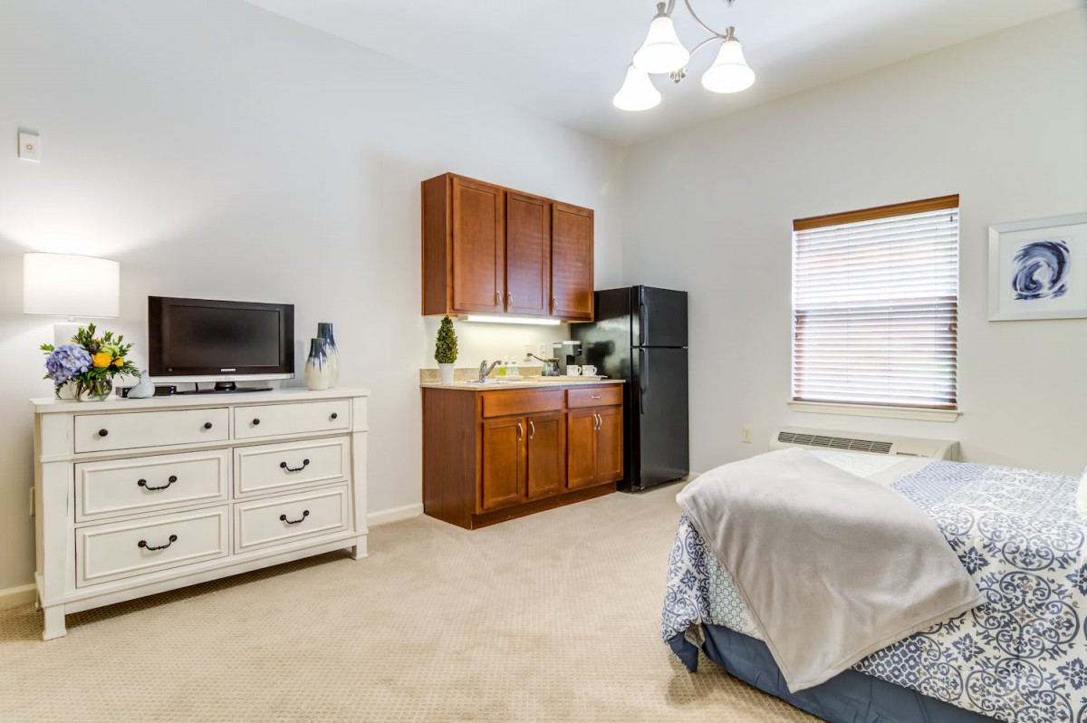 A minimalist bedroom featuring a bed, dresser with a television, and wooden cabinets with appliances.