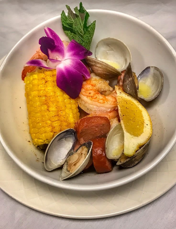 A beautifully plated seafood dish featuring shrimp, clams, corn on the cob, and a lemon wedge, garnished with a purple flower.
