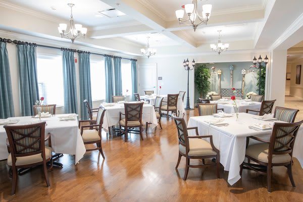 An elegant dining room with neatly arranged tables and chairs, featuring chandeliers and large windows with curtains.