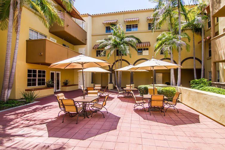 A sunny courtyard with tables and umbrellas surrounded by a yellow building.