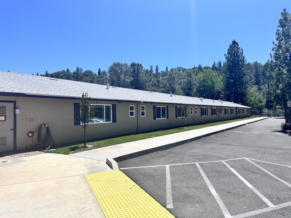 A single-story building with a parking lot and surrounding trees under a clear blue sky.