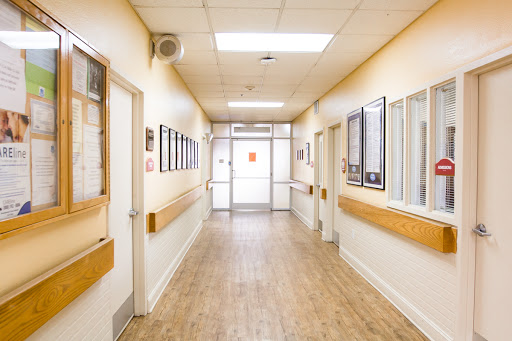 A well-lit hospital hallway, featuring doors and framed images along the walls.