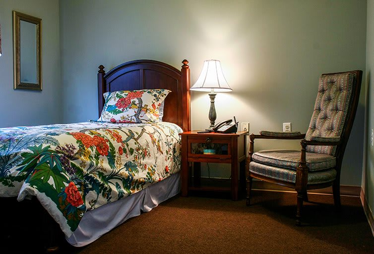 A cozy bedroom with a floral bedspread, wooden nightstand, and chair under a warm lamp light.