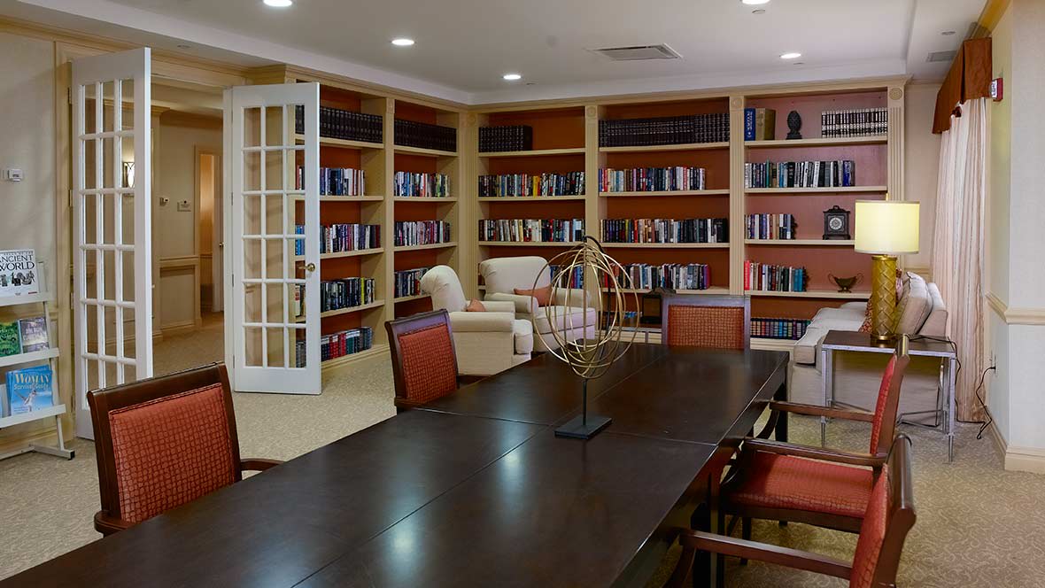 A cozy library room with bookshelves, comfortable chairs, and a large table for reading or meetings.