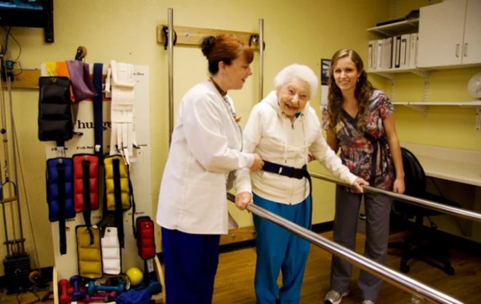 An elderly person receiving support from healthcare professionals during a physical therapy session.