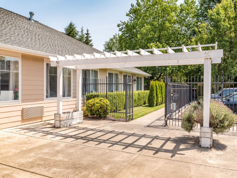 A residential home with a white pergola over the driveway, surrounded by a well-maintained garden.