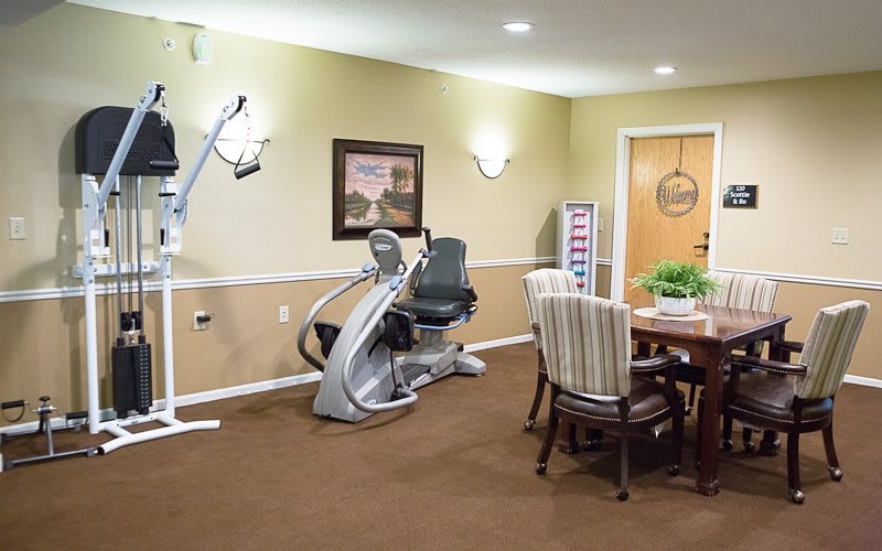 A well-lit fitness room featuring a recumbent exercise bike and a seating area with a table and chairs.