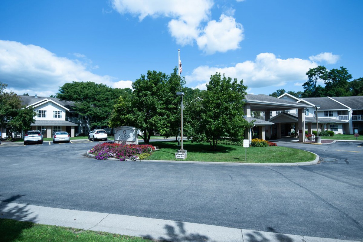 A quiet suburban street lined with trees and residential houses under a partly cloudy sky.