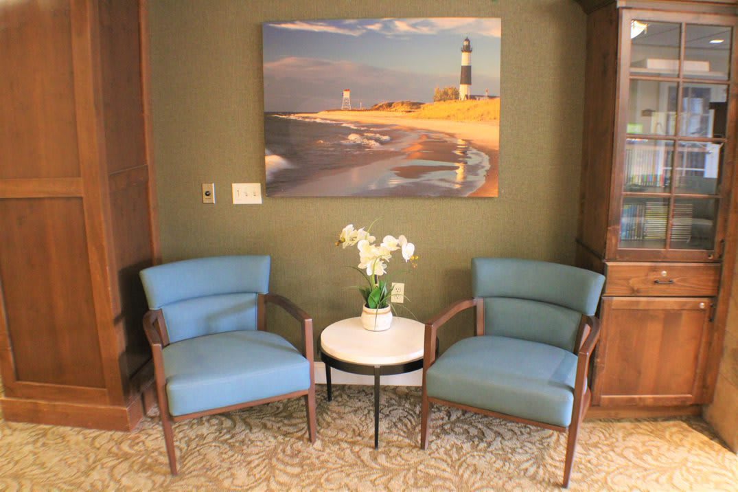 A cozy seating area with two blue chairs and a small round table, adorned with white flowers. A painting of a lighthouse hangs on the wall.