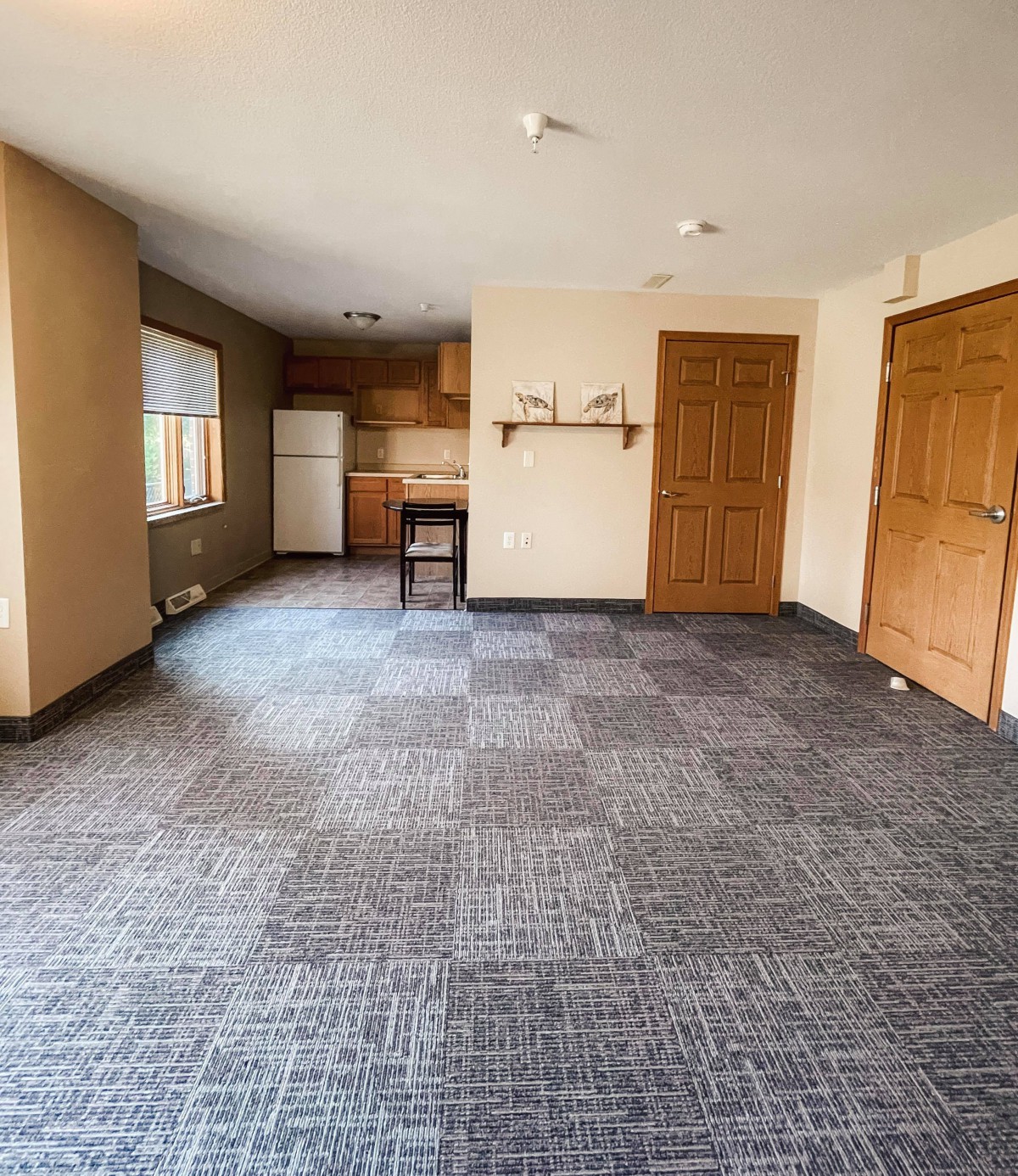 An empty apartment with an open floor plan, featuring a kitchen area and carpeted flooring in neutral tones.