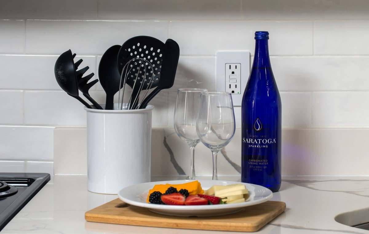 A kitchen setup featuring utensils, a blue wine bottle, glasses, and a plate of fruit on a cutting board.