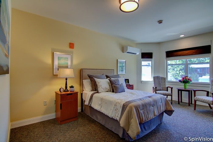A cozy bedroom with a neatly made bed, bedside lamp, and windows letting in natural light.