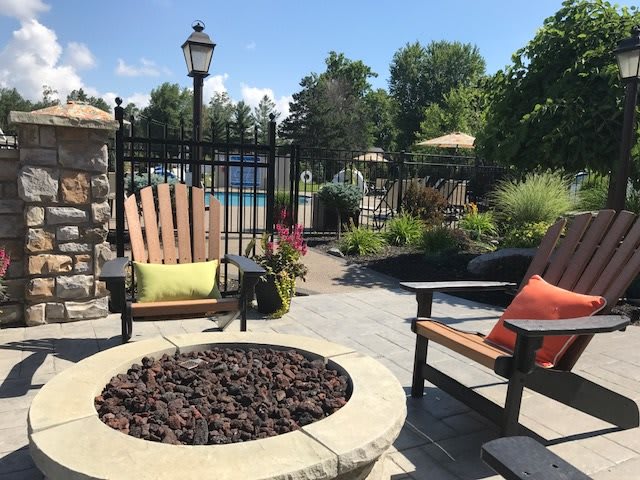 A cozy outdoor patio with a fire pit, surrounded by chairs and lush greenery, near a pool on a sunny day.